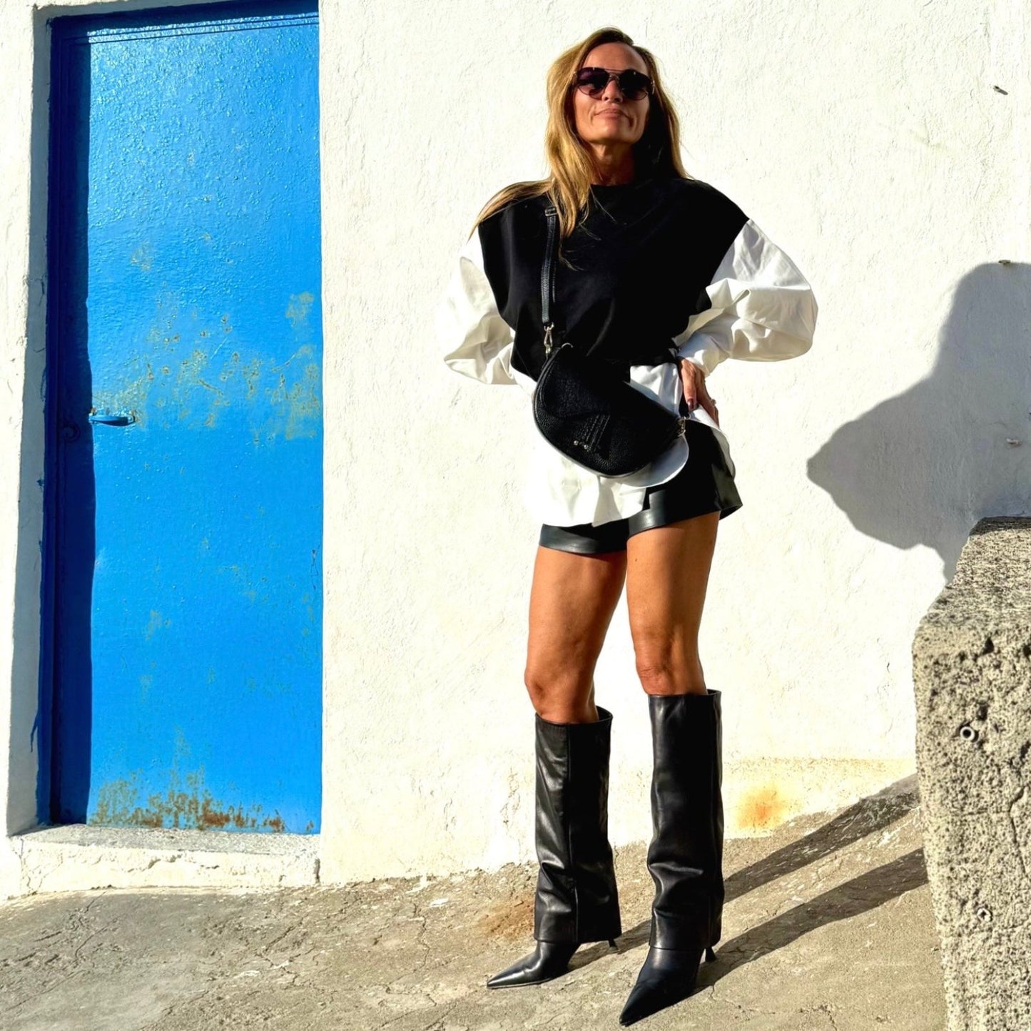 Chloe Boots – LiBEAU Damen trägt schwarze Leder Kniestiefel zu Leder-Shorts, weißem Hemd und schwarzer Weste vor blauer Tür auf Mykonos, selbstbewusster mediterraner Editorial Look | LiBEAU