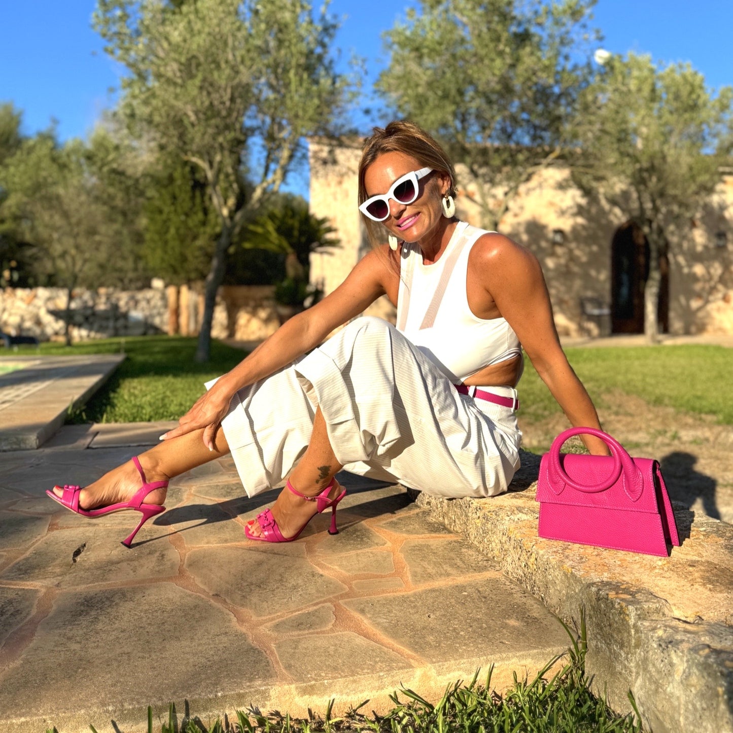 Clutch Viola – LiBEAU Damen sitzt mit Fuchsia Leder Clutch neben sich auf Steinmauer zu weißem Cut-Out-Kleid und Fuchsia-Sandalen in sonniger Mallorca-Finca, strahlender luxuriöser Sommer Editorial Look | LiBEAU
