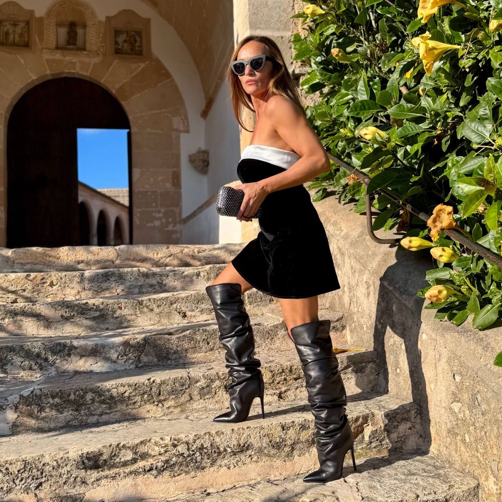Leslie Boots – LiBEAU Damen trägt schwarze Leder Overknee Stiefel zu schwarzem Bandeau-Minikleid und schwarzer Clutch auf sizilianischer Steintreppe, verführerischer luxuriöser Sommer Editorial Look | LiBEAU
