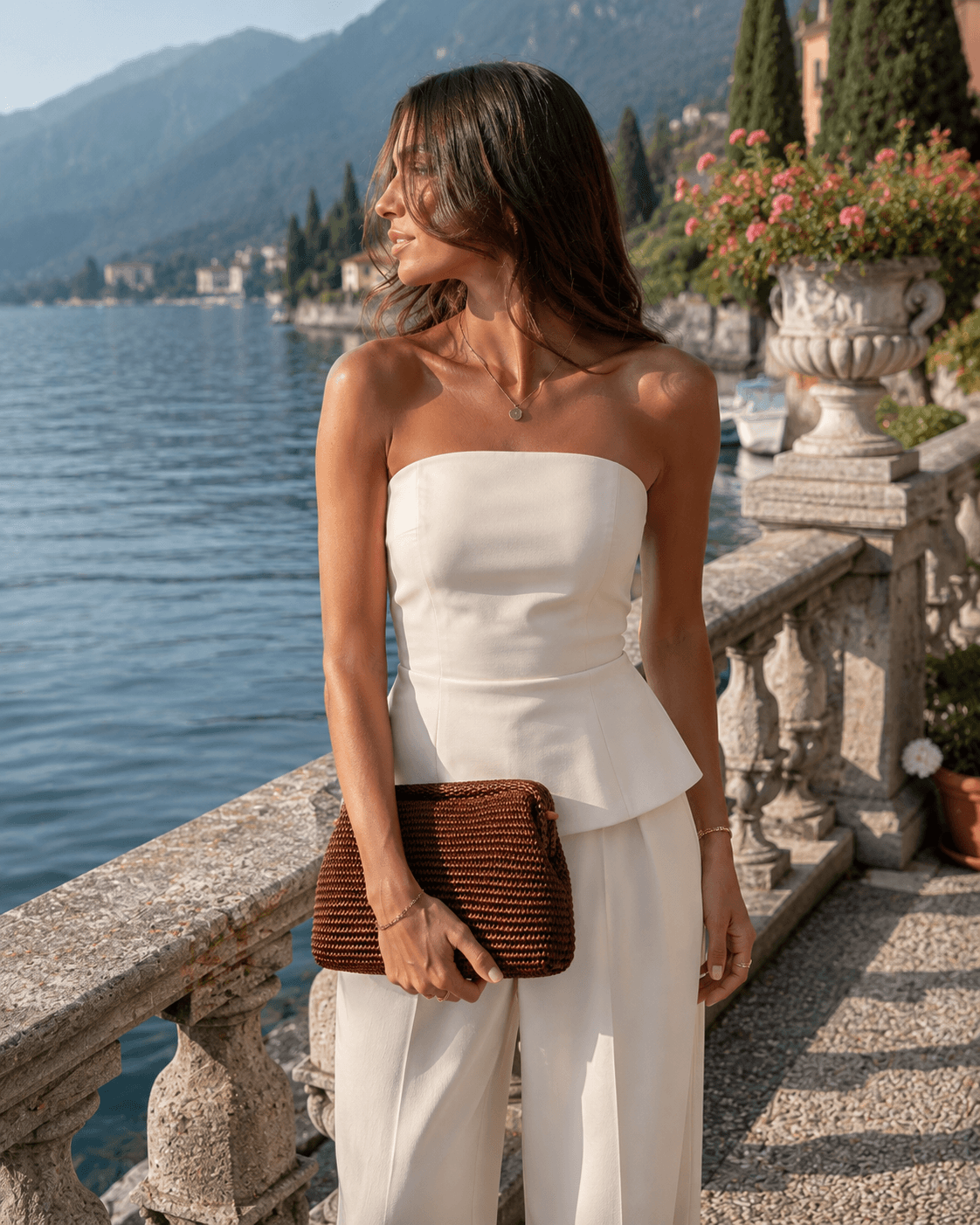 Clutch Rafia Brownie – LiBEAU Damen hält braune Raffia-Clutch zu weißem Bandeau-Jumpsuit und Goldkette auf der Terrasse einer Villa am Comer See, zeitloser luxuriöser Sommer Editorial Look | LiBEAU
