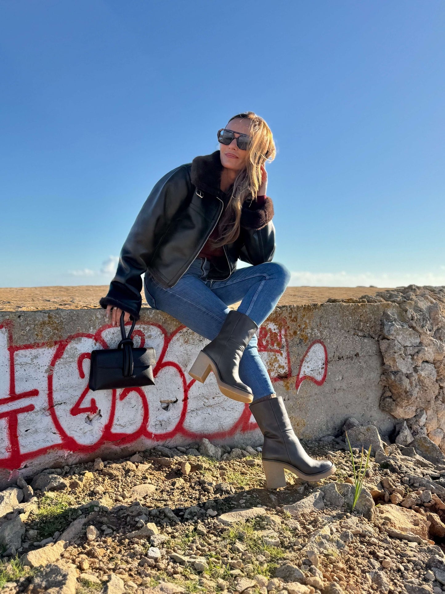 Terra Boots – LiBEAU Damen trägt Dunkelbraun Leder Stiefeletten zu Jeans und Shearling-Lederjacke auf Graffiti-Mauer in Wüstenlandschaft, kraftvoller abenteuerlicher Editorial Look | LiBEAU
